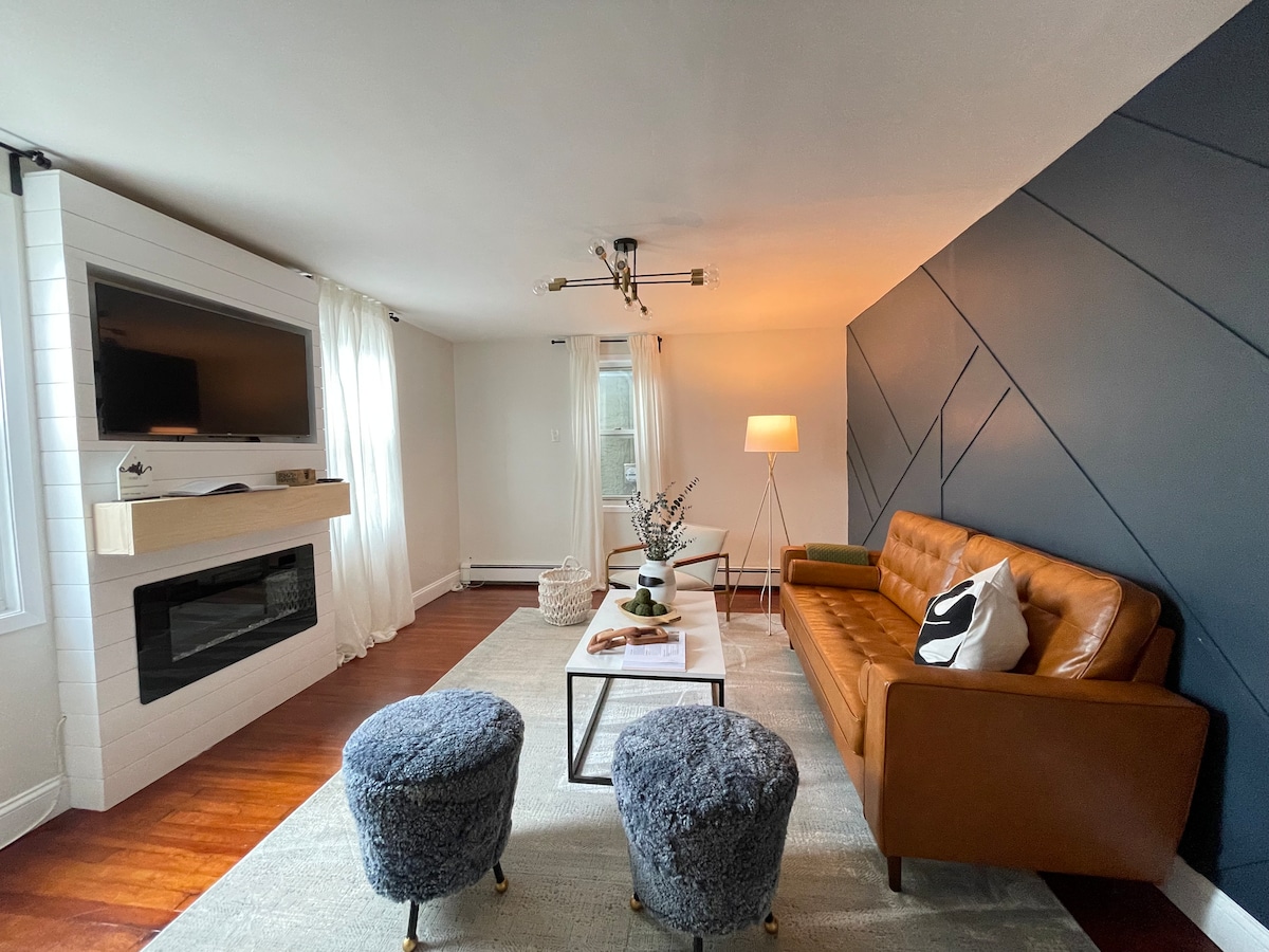 The living room features a stylish leather sofa paired with two plush ottomans. A mounted television and a built-in electric fireplace are positioned on the opposite wall. Natural light filters through sheer curtains, and a coffee table sits atop a soft area rug.
