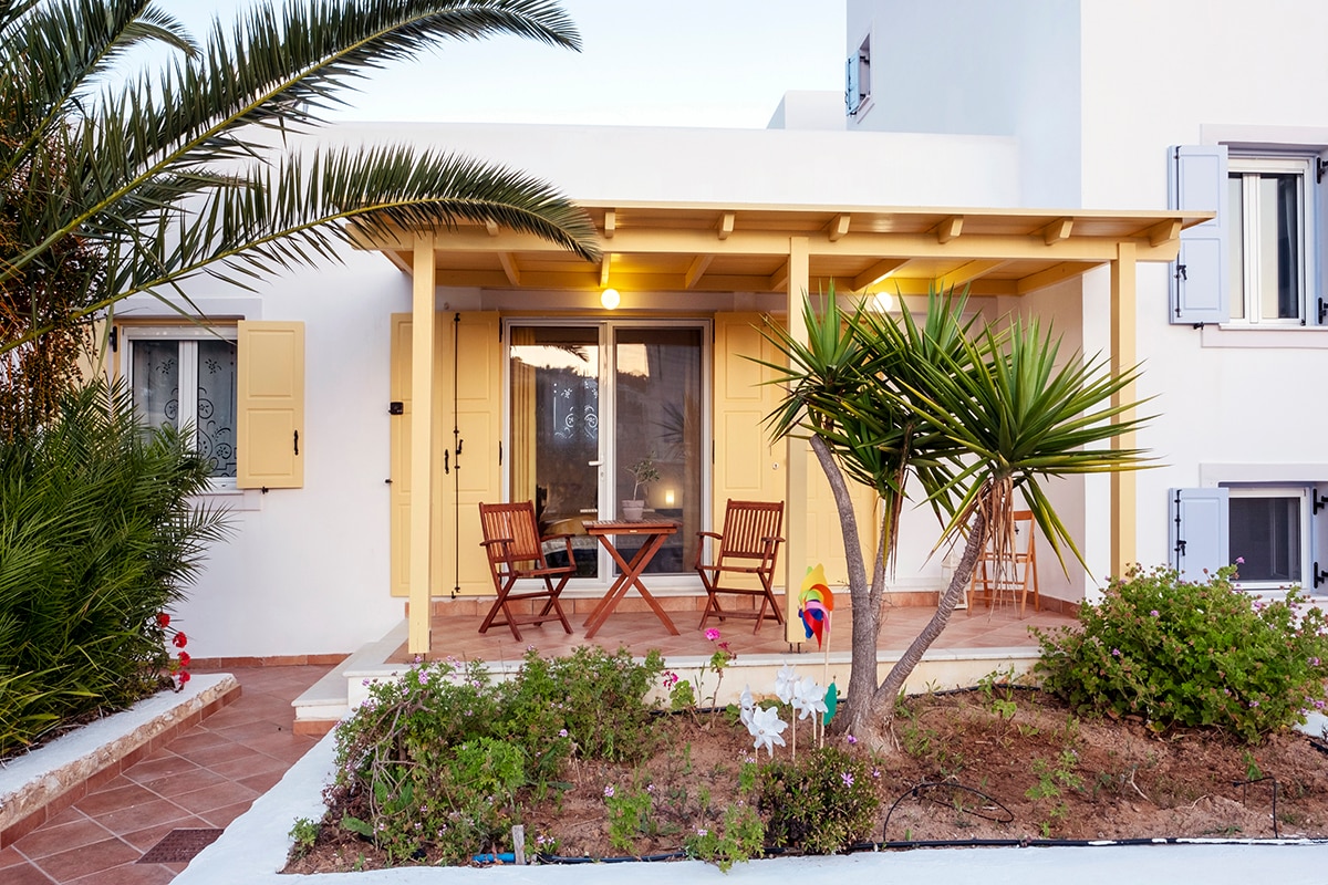 A charming exterior view of the yellow studio presents a covered veranda, furnished with a small table and two wooden chairs. Lush greenery and flowering plants surround the patio, while decorative accents enhance the space. The inviting entrance is framed by bright yellow shutters.