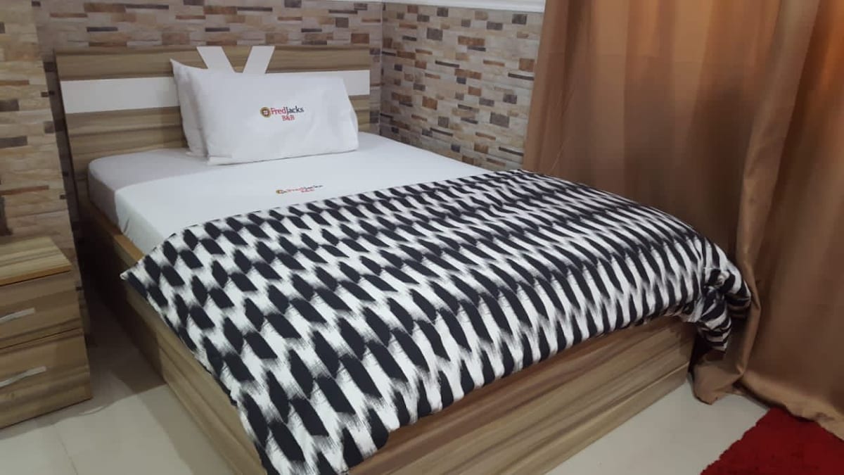 A neatly made bed is positioned against a wall featuring a stone design. The bed is adorned with a monochrome patterned blanket and accented by a decorative pillow. Soft curtains frame the window, allowing natural light to enter the space.