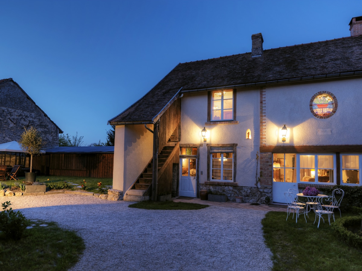 The exterior of a historic home is illuminated by softly glowing lanterns in the twilight. A gravel pathway leads to the entrance, flanked by garden seating and greenery, with the structure showcasing rustic charm and inviting warm light from the windows.