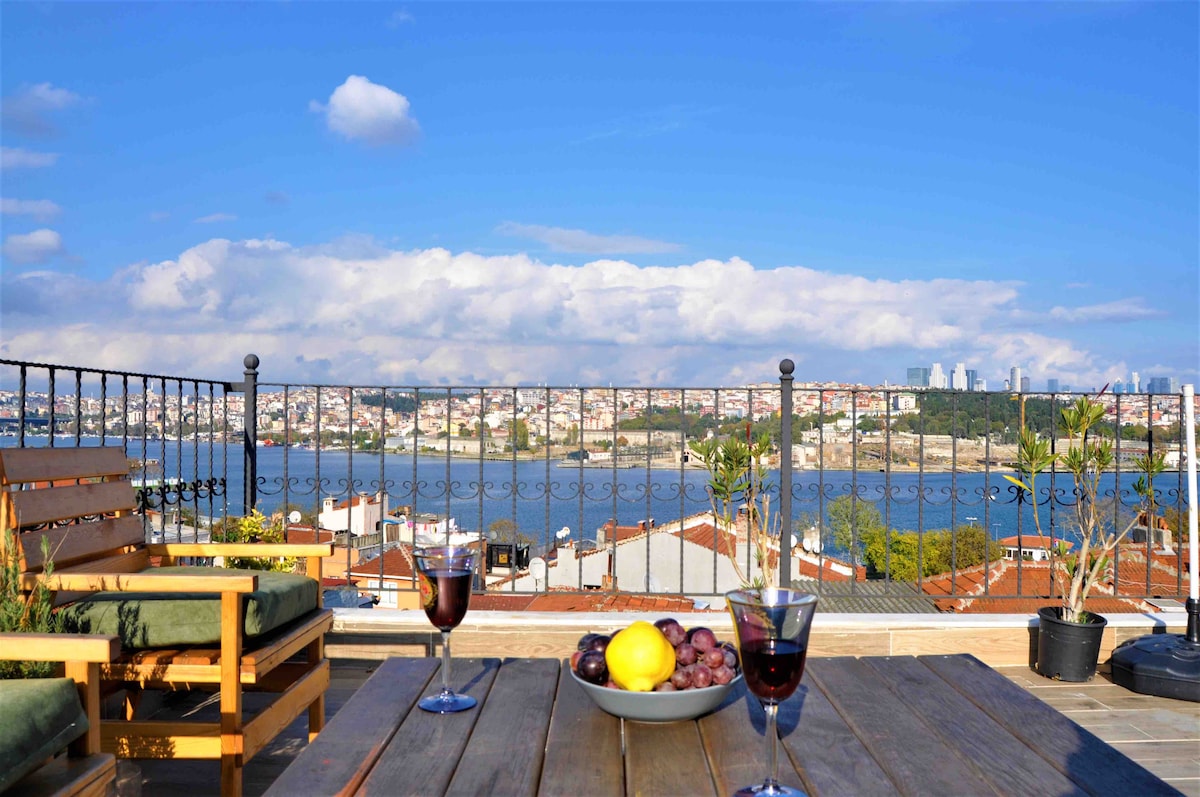 A rooftop terrace offers expansive views of the city and the waterfront. Comfortable seating is arranged around a wooden table, which displays a bowl of fruit and two glasses of dark liquid. The skyline is dotted with buildings beneath a bright, blue sky.
