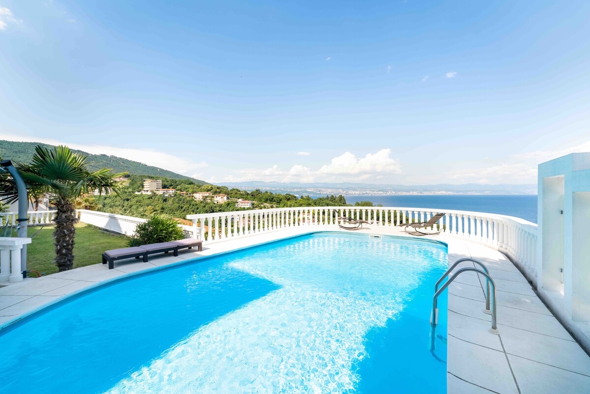 An outdoor swimming pool reflects the clear blue sky, surrounded by a white terrace with railing. Comfortable loungers are positioned for relaxation, and lush greenery can be seen in the background, along with a view of the sea beyond.