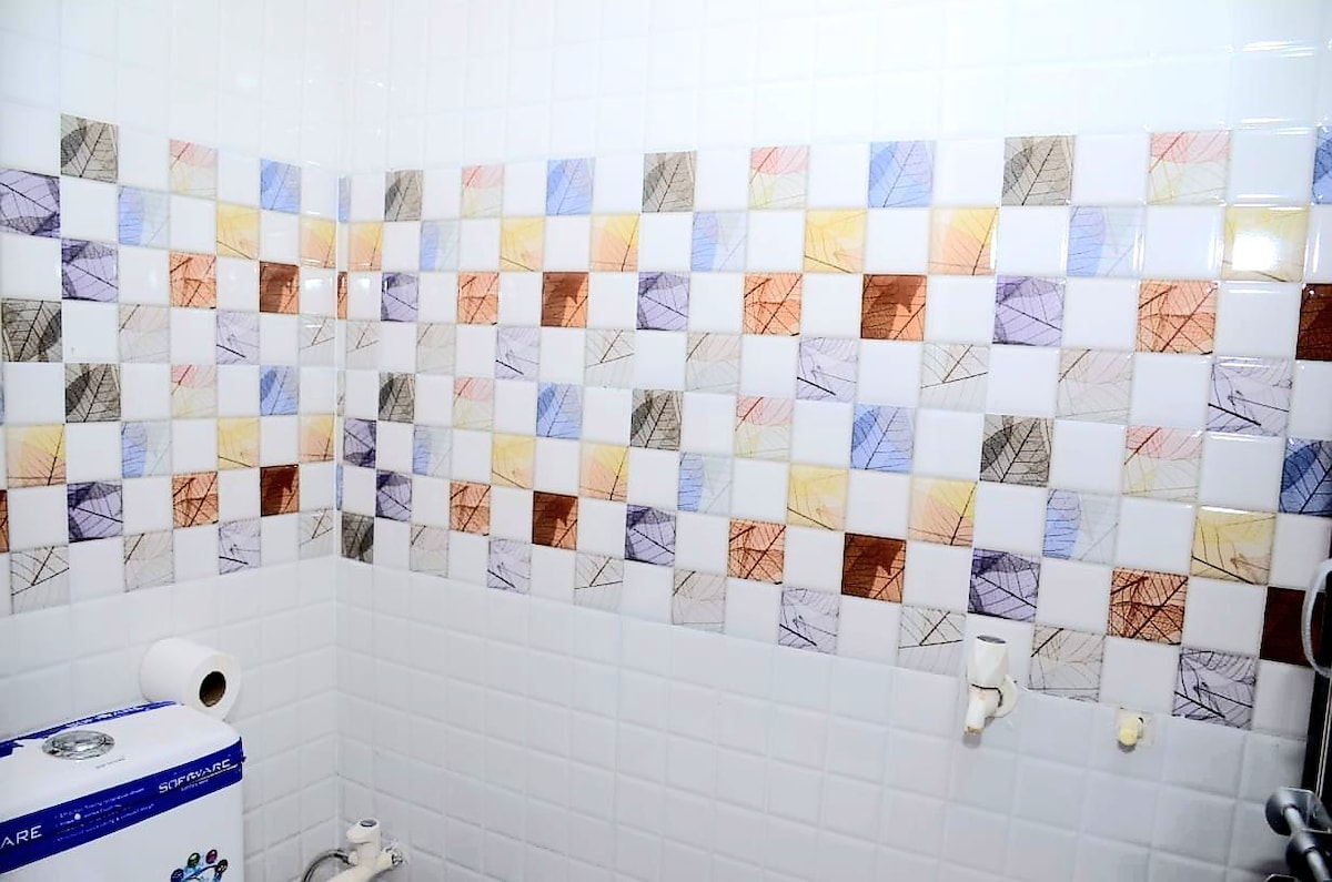 A tiled bathroom wall displays a mosaic pattern of colorful squares in shades of yellow, orange, blue, brown, and gray. The tiles are arranged in a grid format, creating a modern and vibrant aesthetic. A roll of toilet paper and plumbing fixtures are partially visible.
