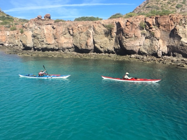 Two kayakers navigate the calm, turquoise waters close to unique rock formations along the shoreline. The rugged rocks rise above the water, creating a striking contrast against the vibrant sea, with gentle waves lapping at the shore.