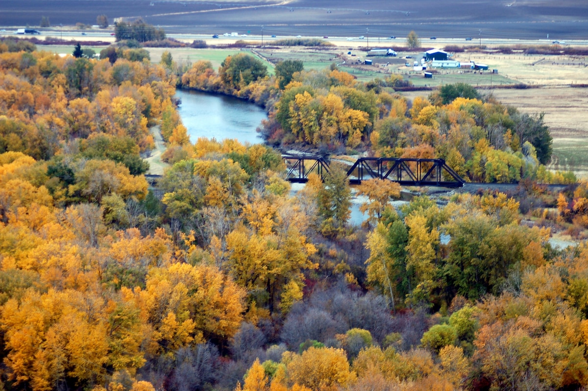 A scenic view displays a winding river surrounded by vibrant autumn foliage. An old railroad bridge spans the river, creating a focal point. The landscape includes fields and structures in the distance, contributing to a sense of rural serenity.
