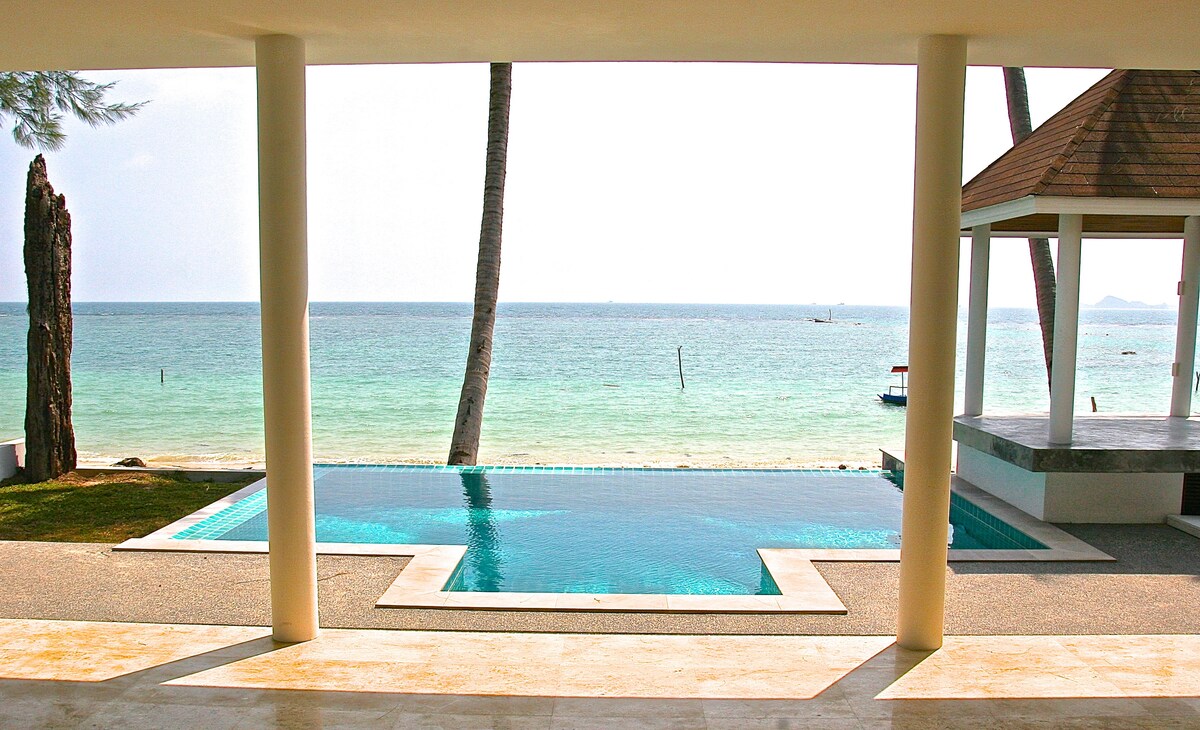 A private swimming pool is visible from the covered patio area, with a clear view of the calm sea beyond. Lush palm trees frame the scene, enhancing the tropical ambiance while sunlight reflects off the water's surface.