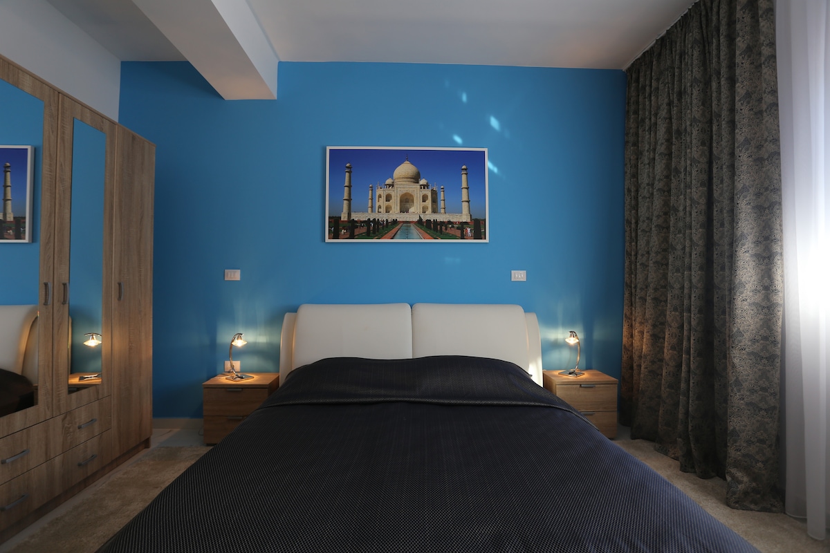 A spacious sleeping area features a comfortable bed with a dark-colored blanket. Two bedside lamps provide soft lighting. A large wardrobe stands against a vivid blue wall, complemented by a striking image of the Taj Mahal. Soft drapes frame the window, adding an elegant touch.
