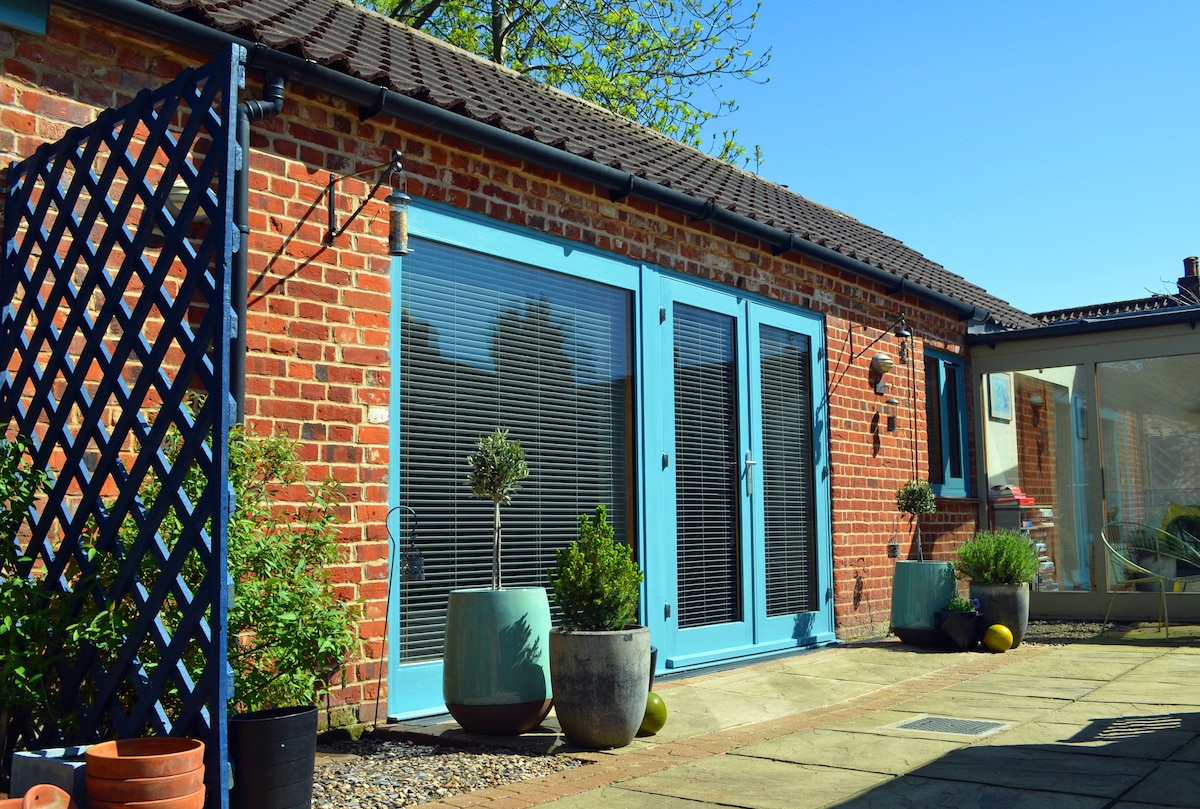 The exterior of the guest suite features double glazed doors framed by vibrant blue accents. Potted plants in varying sizes are positioned on either side of the entrance. A courtyard of neatly arranged paving stones leads up to the suite, illuminated by natural sunlight.