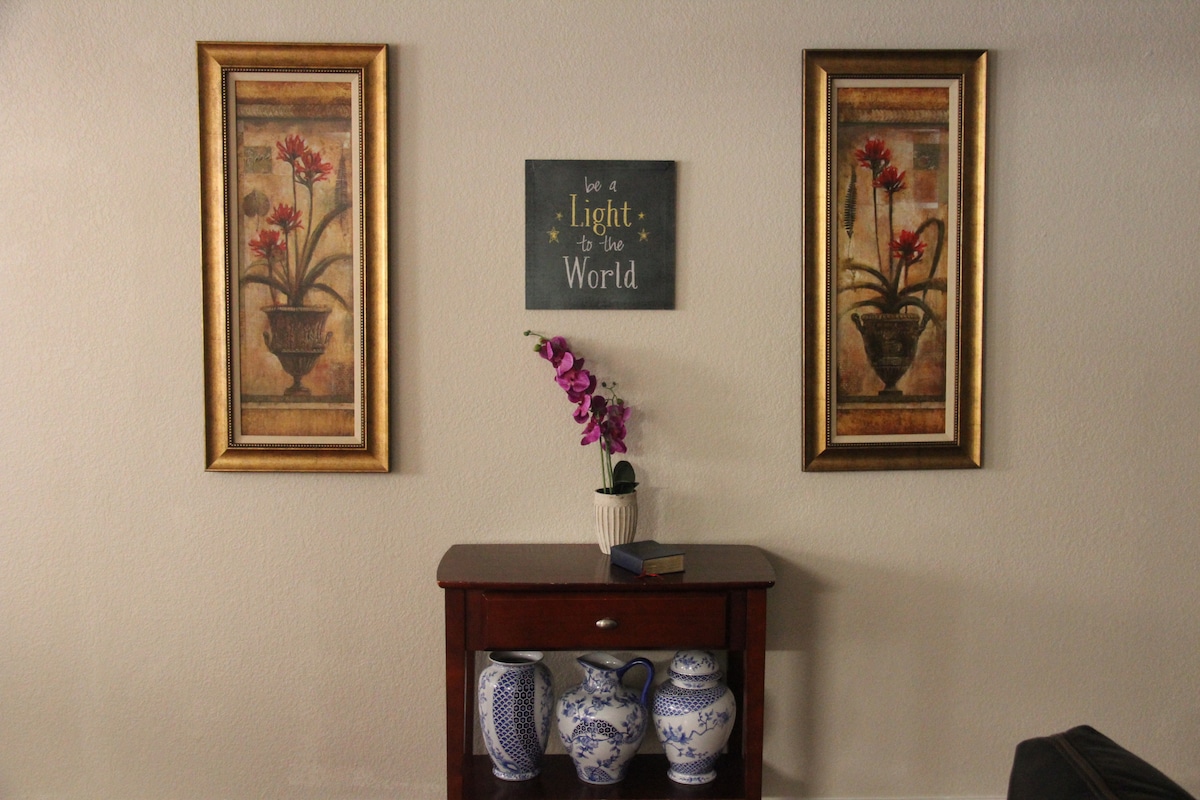 A small wooden table is positioned against the wall, adorned with blue and white ceramic vases. Framed floral artwork hangs on either side, creating a balanced aesthetic. A decorative sign with the phrase 'be a Light to the World' is displayed above the table, adding to the inviting ambiance.
