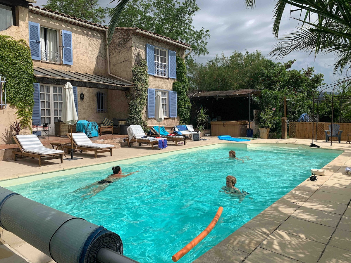 A spacious outdoor pool area is surrounded by sun loungers and umbrellas. The villa, featuring blue-shuttered windows, is visible in the background, along with lush greenery. A covered patio area is present, providing additional shade and seating options.