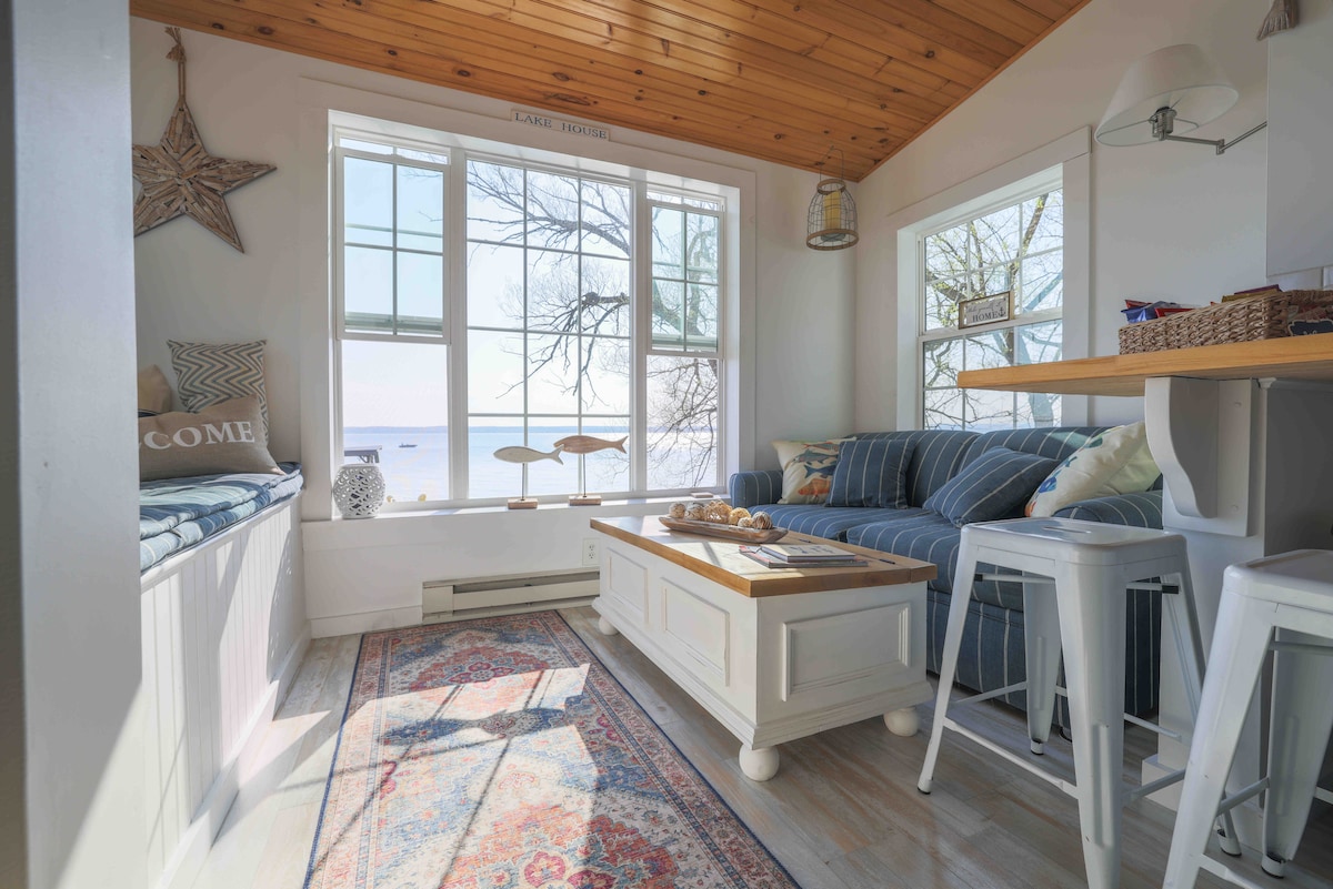 A bright living space features a cozy blue-striped sofa and a coffee table set atop a decorative area rug. Large windows offer beautiful views of the lake, while natural light fills the room. Additional seating is provided by white stools at the nearby table.