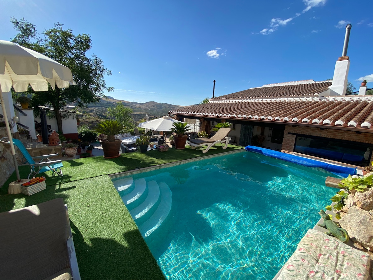 A sunlit outdoor area features a private pool with steps leading into the water. Surrounding the pool are potted plants and lounge chairs. The villa's exterior, with tiled roof and shaded seating areas, blends harmoniously with the scenic hill views in the background.