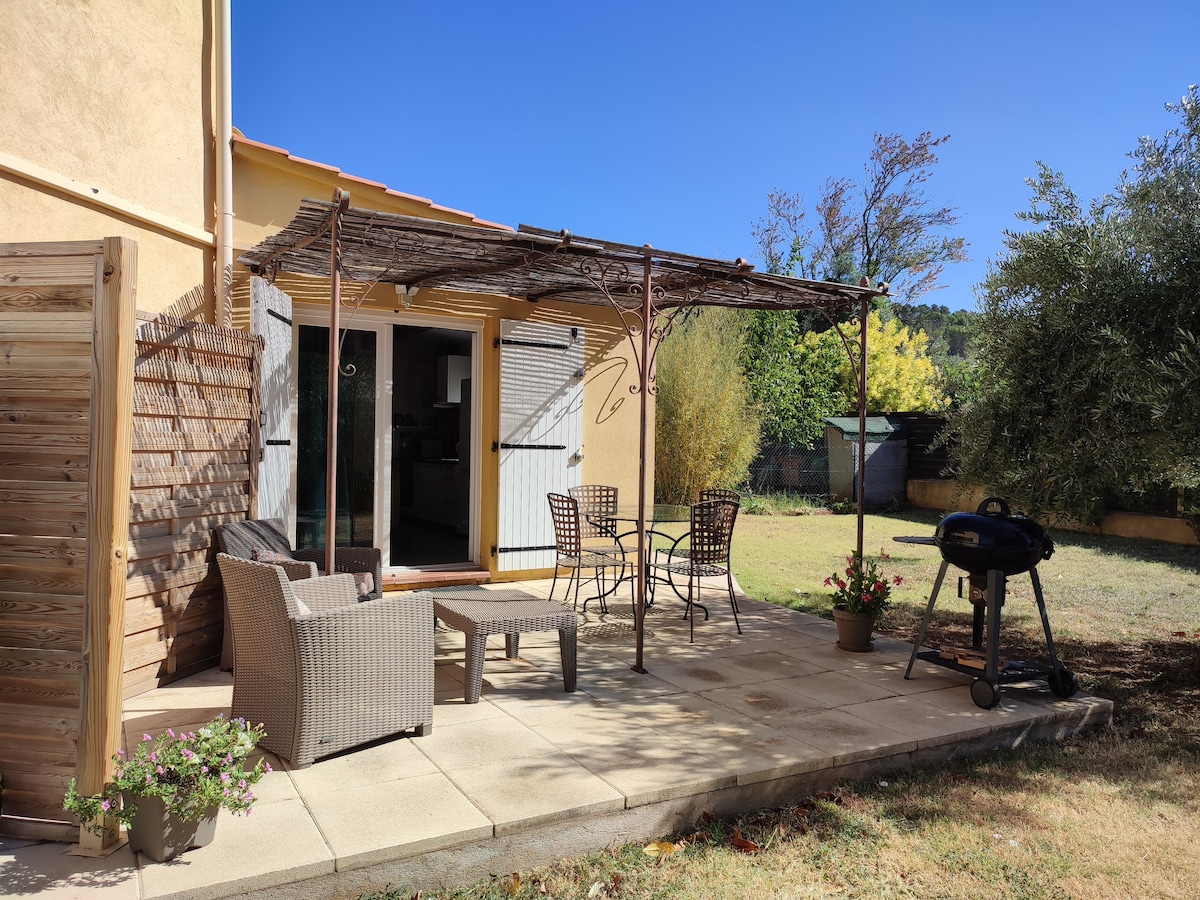 A covered terrace is featured, furnished with a glass-top table and four chairs, surrounded by potted plants. A barbecue grill sits nearby on a tiled surface, with views of the well-maintained lawn and surrounding greenery.