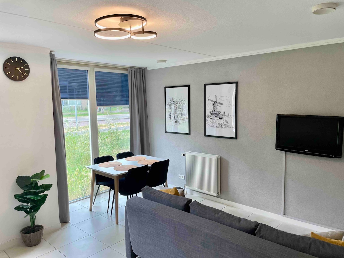 The open living area features a comfortable gray sofa facing a wall-mounted TV. A dining table with four black chairs is positioned beside large windows with sheer curtains. Two framed art pieces and a clock complement the light gray walls. A decorative plant adds a touch of greenery.
