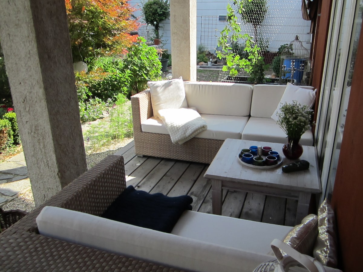 A cozy outdoor seating area is presented, featuring a white sectional sofa and a wooden table. Colorful ceramic dishes are arranged on the table, while greenery and garden elements are visible in the background, creating a relaxing environment for guests.