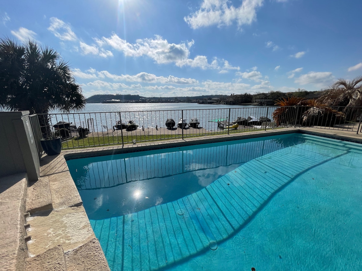 A refreshing pool is shown with clear blue water reflecting the sky. Lounge chairs are visible nearby, positioned along the water's edge, under a sunny sky dotted with clouds. Lush greenery adds a natural backdrop, enhancing the serene lakeside setting.