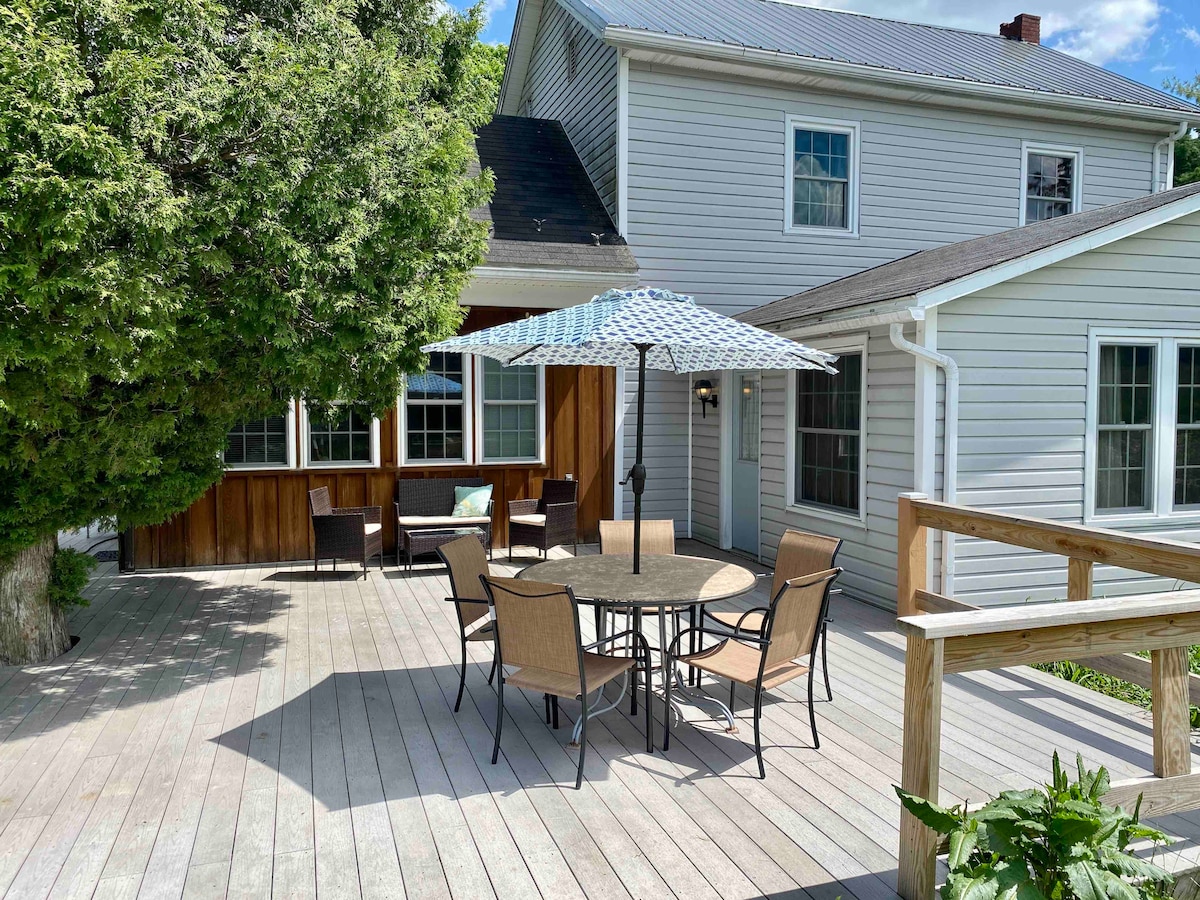 A spacious deck features a round dining table surrounded by six chairs, shaded by an umbrella. Cozy seating areas are arranged nearby, offering a comfortable space for relaxation. The white farmhouse exterior is complemented by lush greenery and large windows allowing ample natural light.