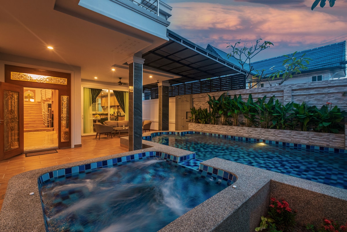 A private swimming pool and jacuzzi are set against a serene backdrop, surrounded by lush greenery. The area is bathed in soft evening light, showcasing a modern aesthetic that invites relaxation. The entrance to the villa is visible, with wooden doors leading to the interior.