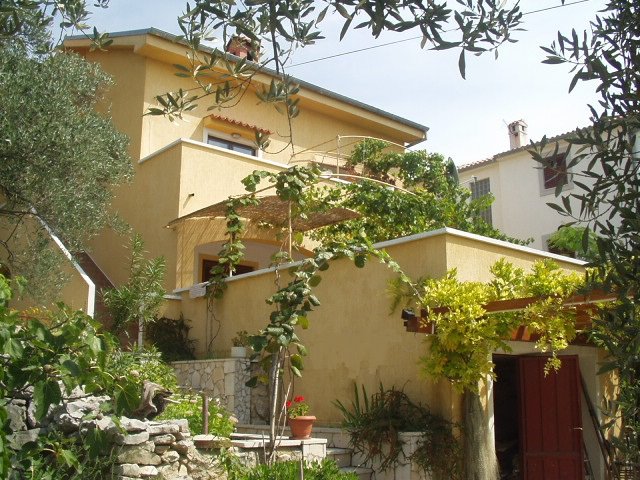 A two-story apartment building is surrounded by lush greenery, including olive trees and climbing vines. A terrace with a shaded area is visible on the upper level, while a rustic wooden door leads to the entrance. The exterior is painted in a warm yellow hue.
