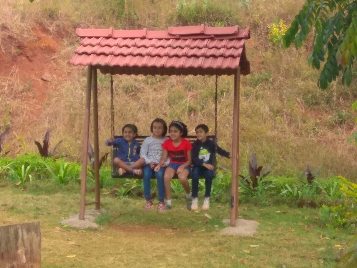 A wooden swing is situated in a grassy area, surrounded by lush greenery. Four children sit on the swing, enjoying the outdoor space which features a rustic roof overhead. The backdrop includes gently sloping hills and a variety of plants, enhancing the serene environment.