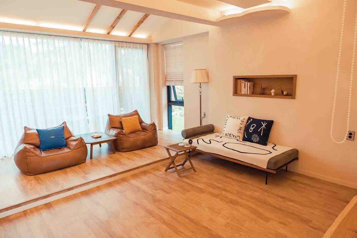 A bright living area is highlighted by large windows covered with sheer curtains, allowing natural light to fill the space. Two bean bags and a simple sofa are arranged comfortably around a small wooden table, providing a relaxed atmosphere. Warm wood flooring enhances the inviting setting.