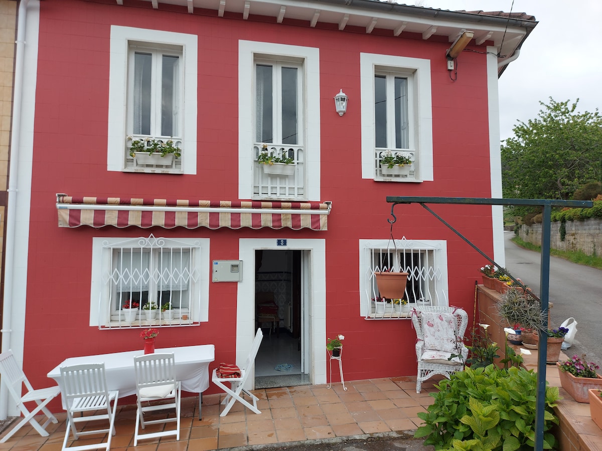 A charming two-story house features vibrant red exterior walls with white window frames. White patio furniture is arranged on the terrace, complemented by potted plants, creating an inviting outdoor area. The entrance is adorned with decorative elements that enhance the welcoming appearance of the home.