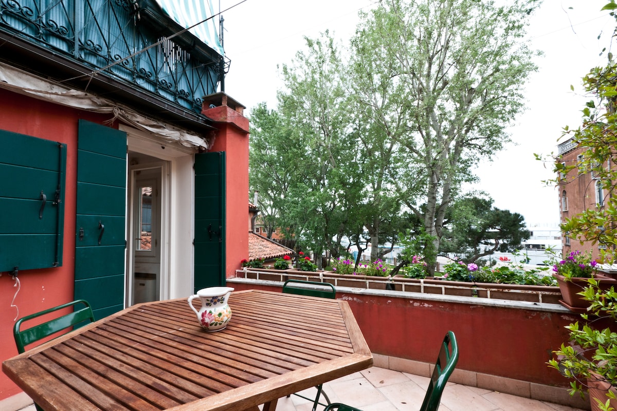 An outdoor terrace features a wooden table surrounded by green chairs, offering a space for dining. Potted plants add touches of greenery, while a large tree nearby provides shade. The terrace is framed by colorful window shutters, creating a pleasant outdoor setting.