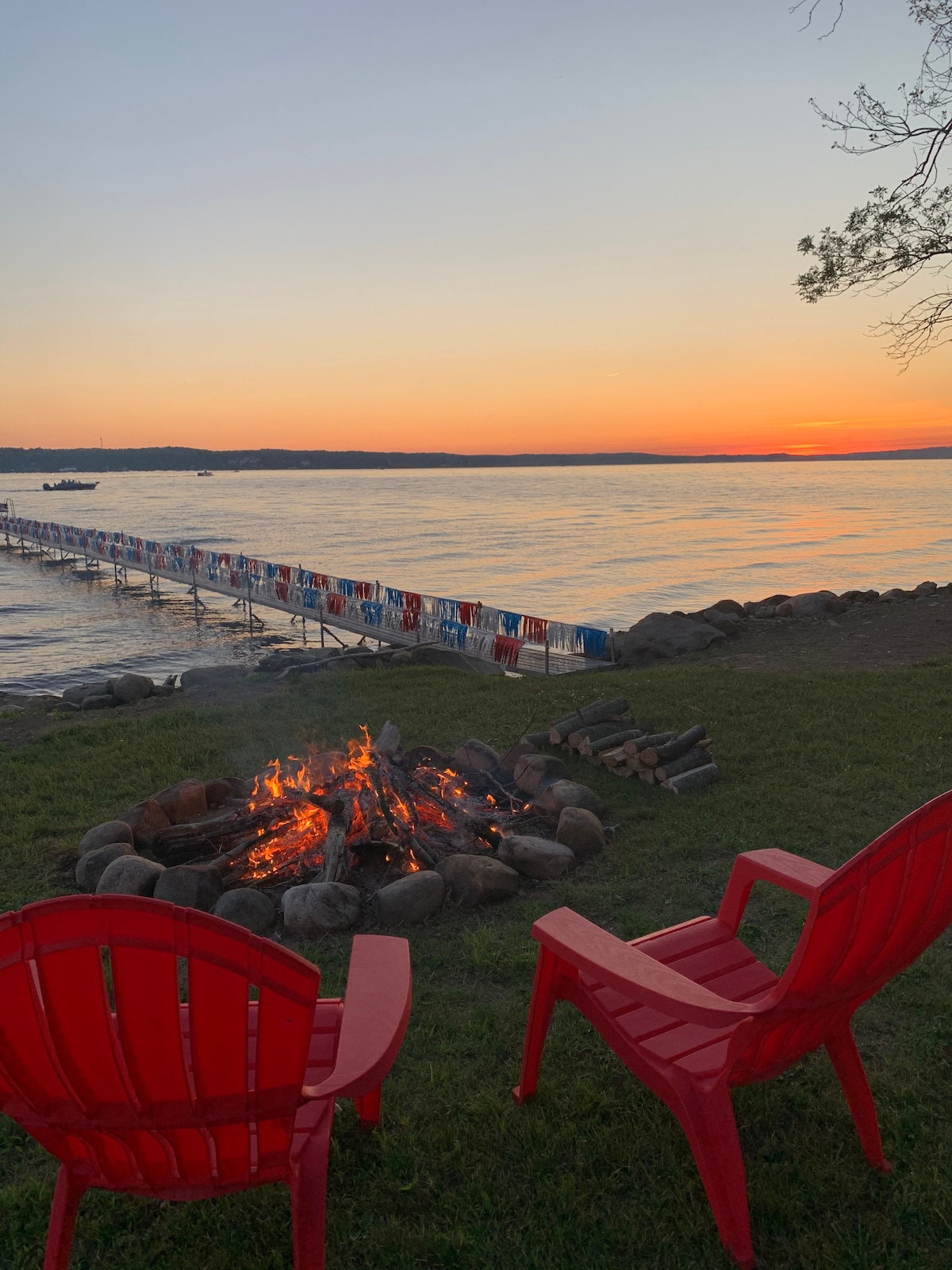 A fire pit surrounded by stones is positioned on a grassy area, facing the water. Two bright red chairs are arranged nearby. In the distance, a wooden dock, adorned with colorful beach towels, extends into the lake, framed by a serene sunset over the horizon.