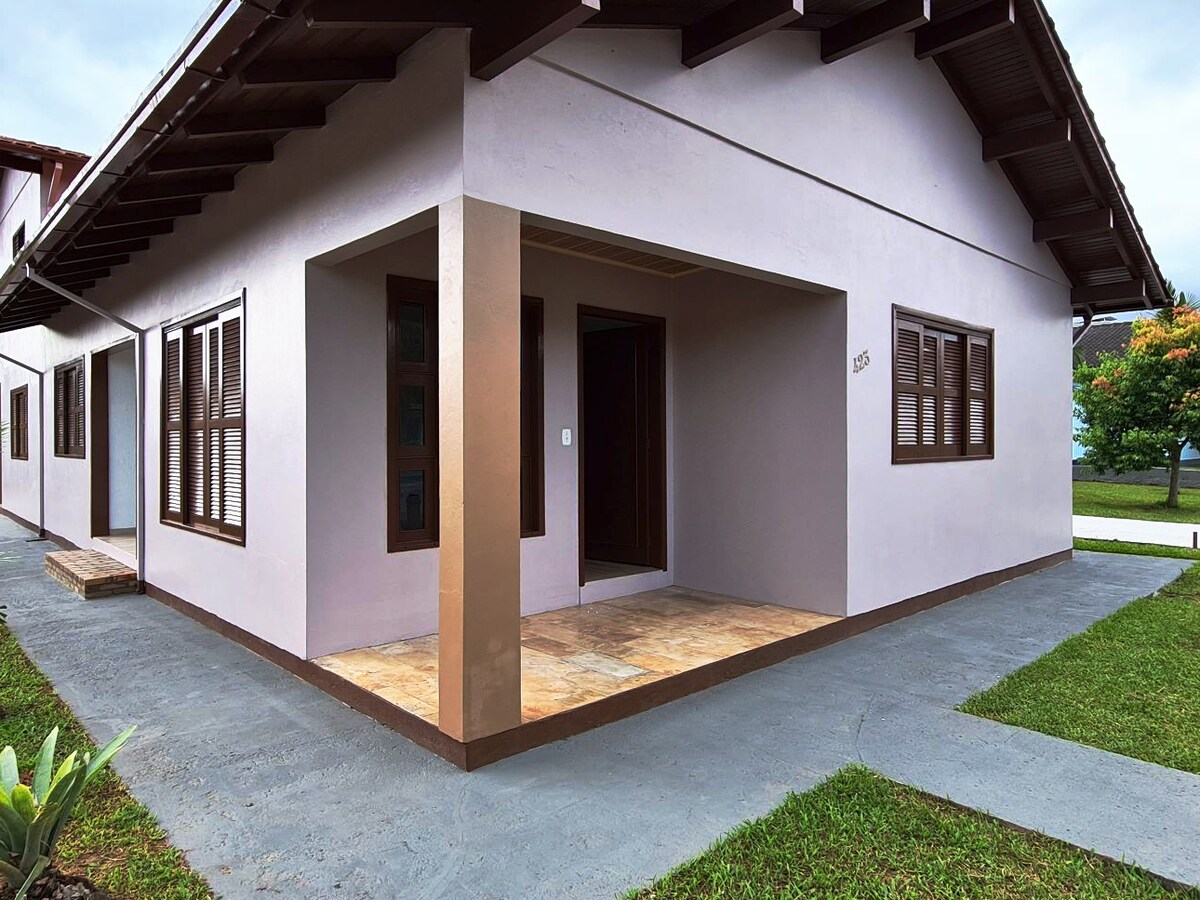 A welcoming entrance is featured, with a spacious porch bordered by light-colored tiles. The exterior walls are painted in soft hues, complemented by wooden window frames. Lush greenery is visible in the surrounding area, contributing to a calm environment.