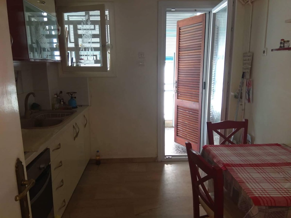 A well-equipped kitchen area features white cabinetry and modern appliances on one side, while a small dining table with red chairs is positioned nearby. A door leads to an outdoor space, allowing natural light to fill the room.