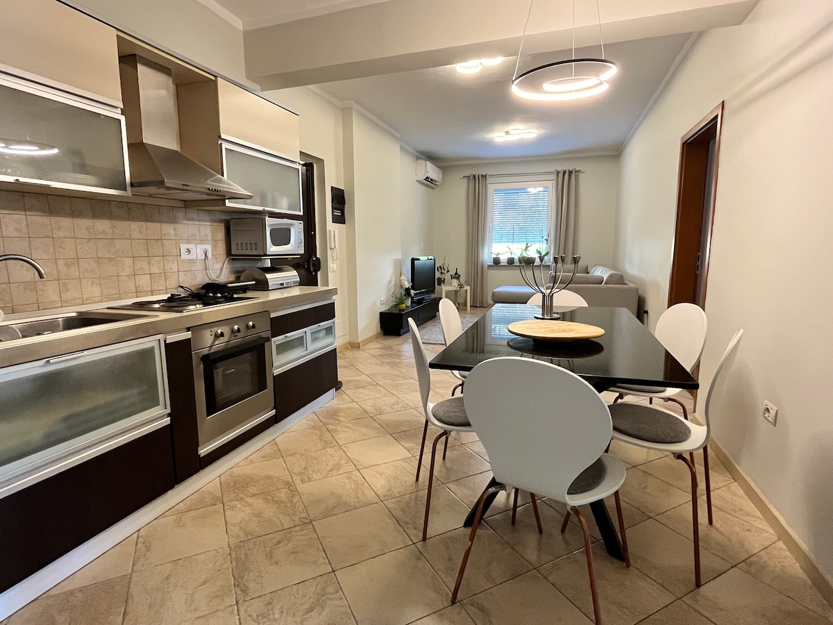A modern kitchen and dining area are shown, featuring a sleek design. Stainless steel appliances are neatly arranged, with a dining table set for four. Natural light filters through a window, illuminating the space's neutral tones and highlighting the adjacent living area.