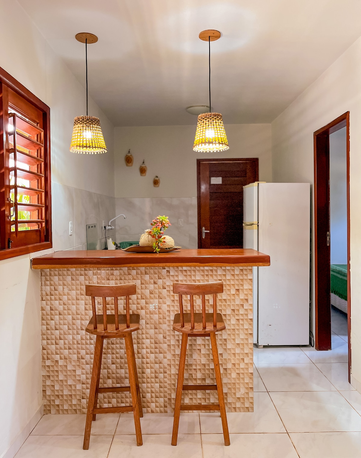 A cozy kitchen area features a bar with two wooden stools and pendant lights. The backsplash is designed with textured tiles. A refrigerator stands against the wall, while subtle decorations are displayed above the sink, creating an inviting space for meal preparation.