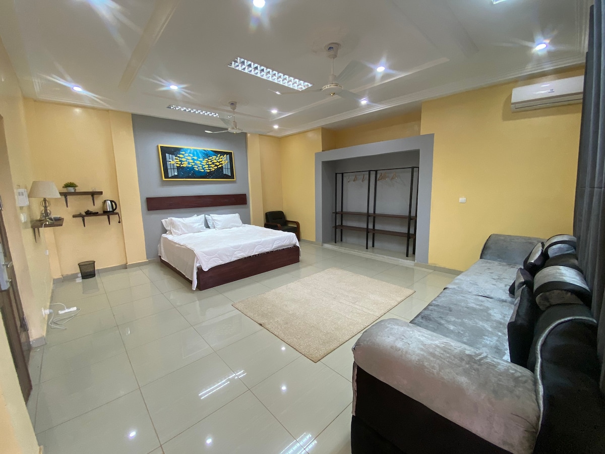 An open living area features a spacious bed, complemented by a soft area rug at its foot. A comfortable sofa is positioned nearby, and minimalist shelving is mounted against the wall. Natural light brightens the room through strategically placed lighting fixtures.