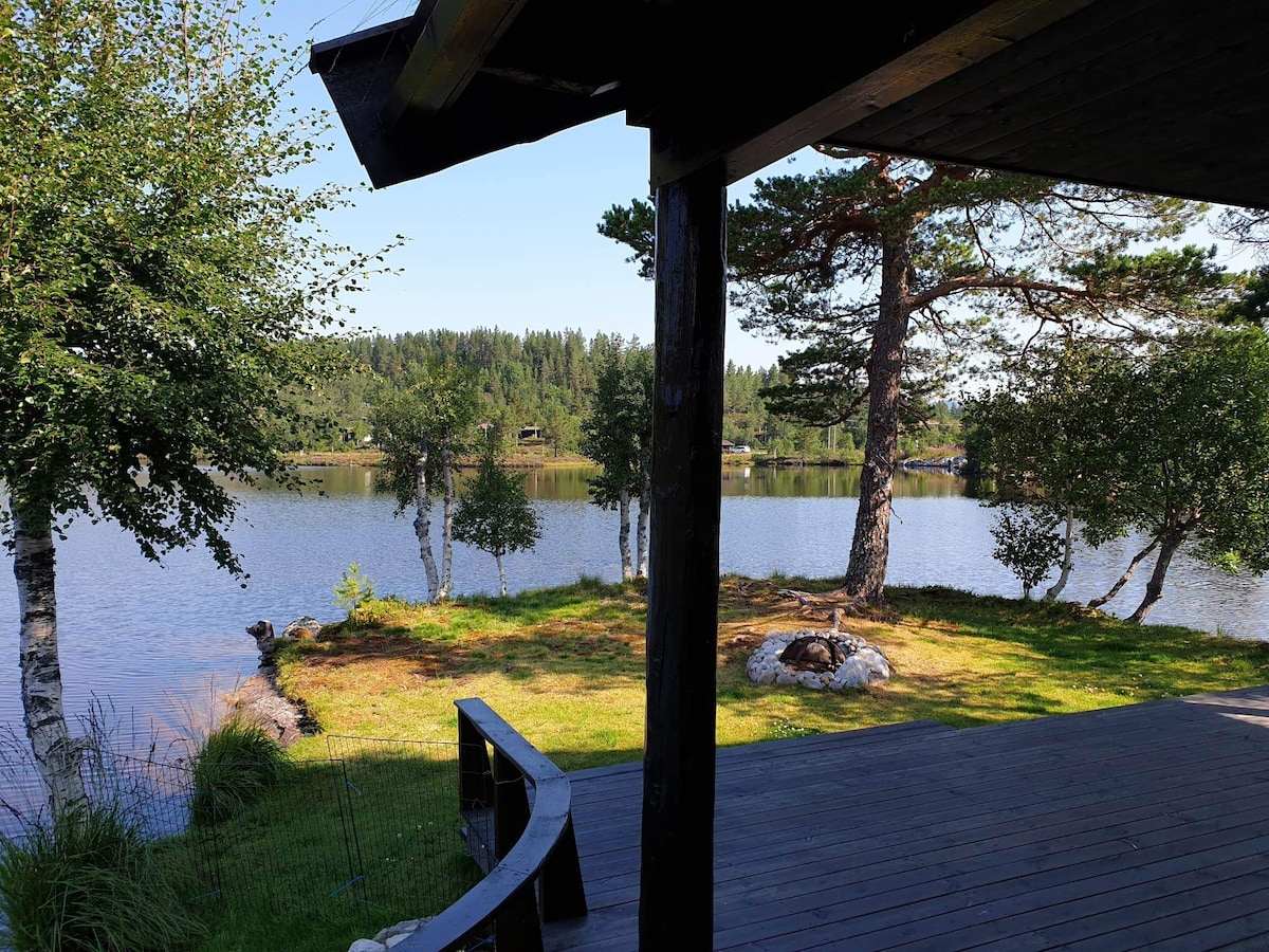 A serene view across a calm lake is framed by trees and a grassy shore. The image captures the peaceful natural landscape, with gentle water reflecting the surrounding greenery and distant hills.