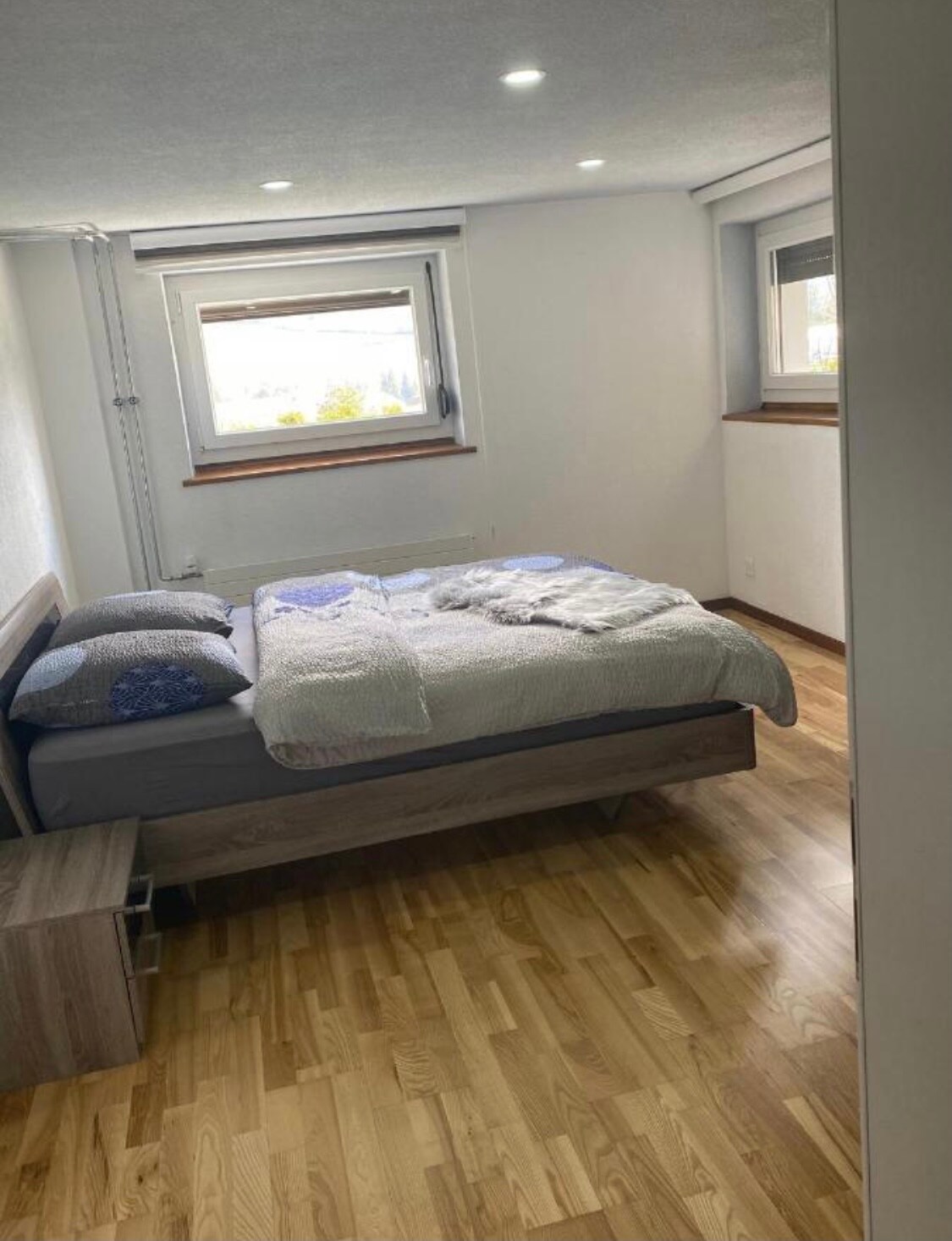 A bedroom featuring a spacious bed dressed in neutral linens with an assortment of pillows. Natural light enters through two large windows, enhancing the wooden flooring. A bedside table is positioned beside the bed, offering a minimalistic design.