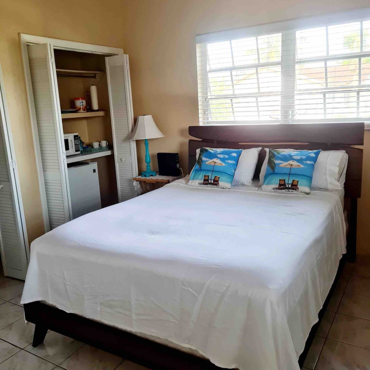 A spacious bedroom features a large bed adorned with decorative pillows. A lamp and a nightstand are beside the bed. A closet contains a mini-fridge and microwave, while natural light enters through the window with white blinds.