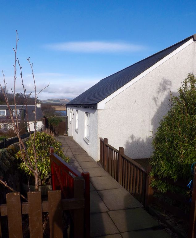 A pathway leading to the entrance of the detached house is flanked by greenery and a wooden fence. The exterior wall features two windows, with sunlight illuminating its white paint, and a clear blue sky serves as a backdrop.