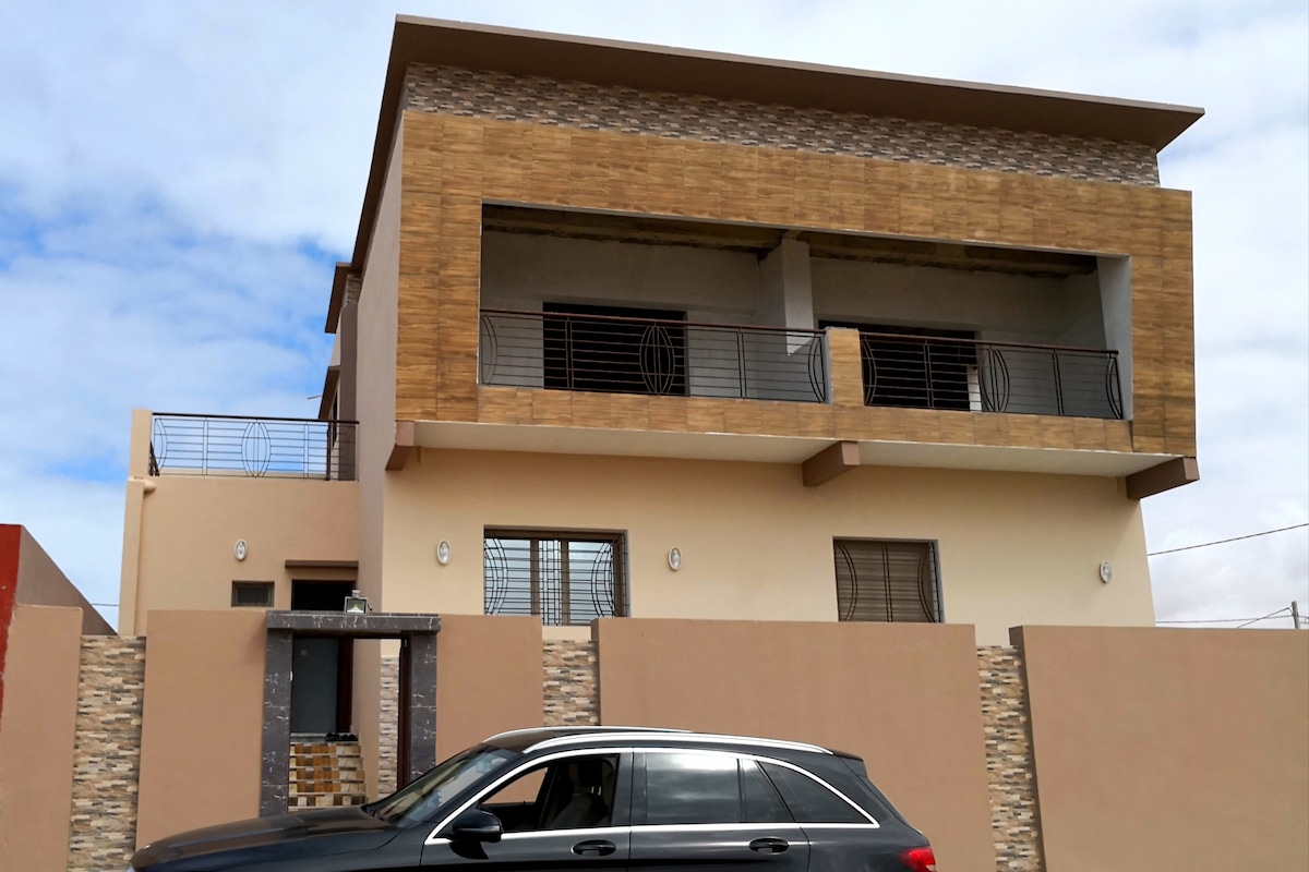 The exterior of a modern villa is showcased, featuring a mix of textured wood and smooth materials. Two spacious balconies are visible on the upper floor, surrounded by decorative railings. A car is parked in front, and a smooth stone wall provides privacy.
