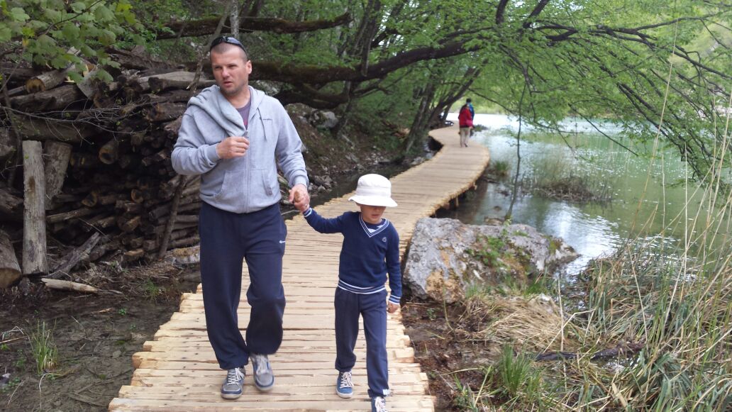 A wooden pathway follows the edge of a serene body of water, framed by lush greenery. Two individuals are seen walking together, with a child holding an adult's hand. In the distance, a person appears to be strolling along the same path.