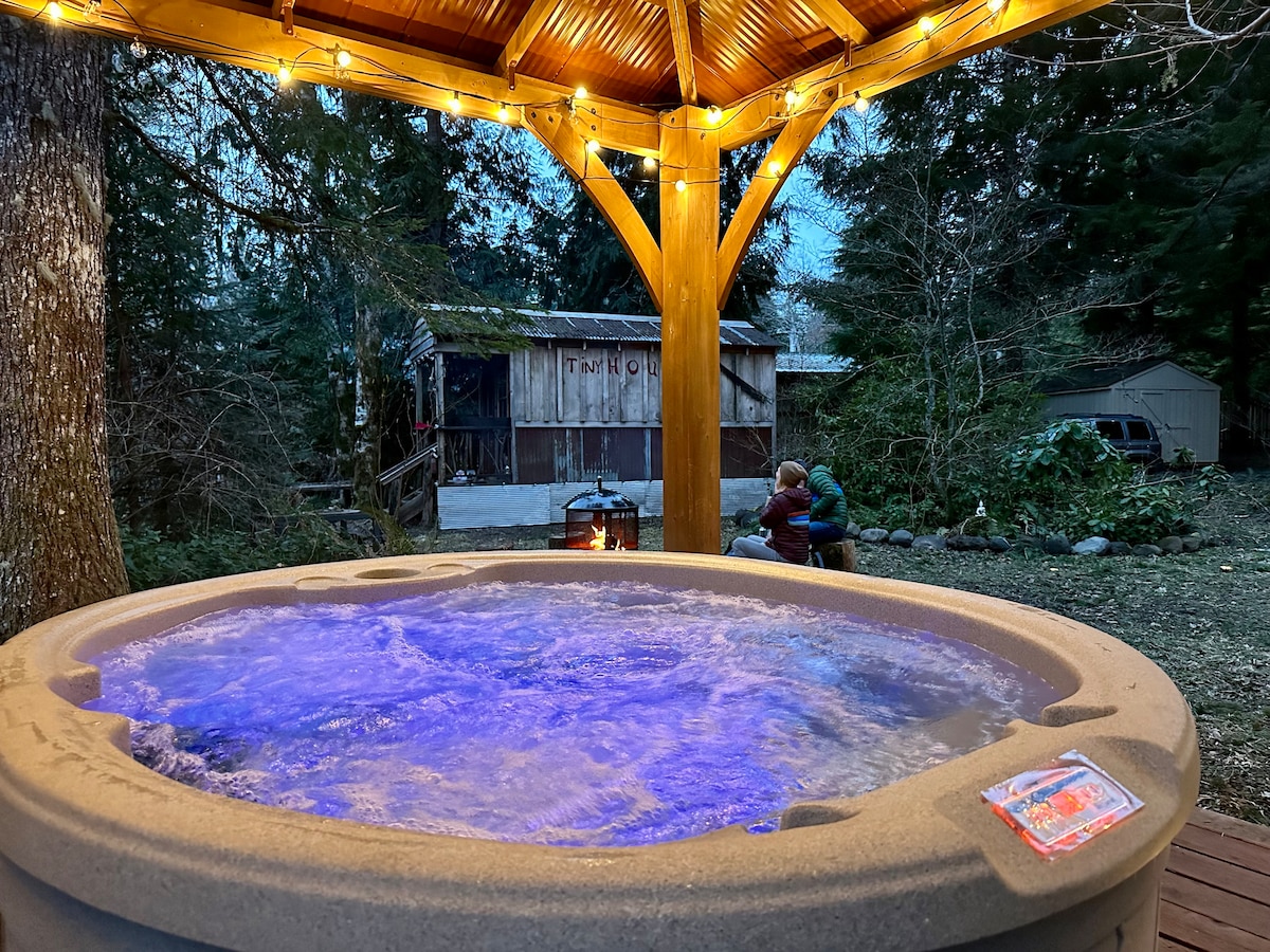 A hot tub is set beneath a wooden gazebo, illuminated with string lights. The water appears inviting with a soft blue hue. In the background, a rustic cabin is partially visible, alongside a fire pit designed for cozy gatherings.