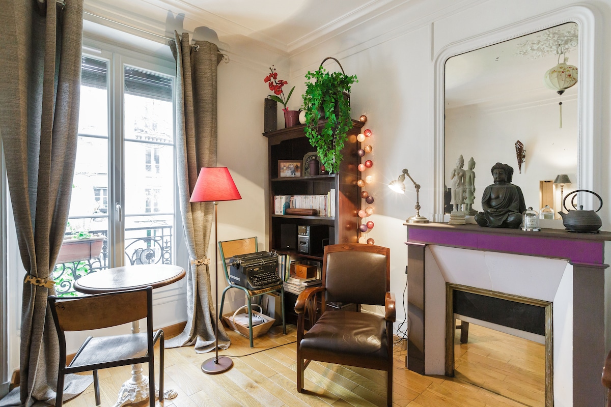 A cozy corner of the living room is shown, featuring a small round table paired with a wooden chair. A comfortable brown leather armchair is positioned nearby, and a bookshelf displays various items. Natural light enters through the large window, adorned with elegant curtains.