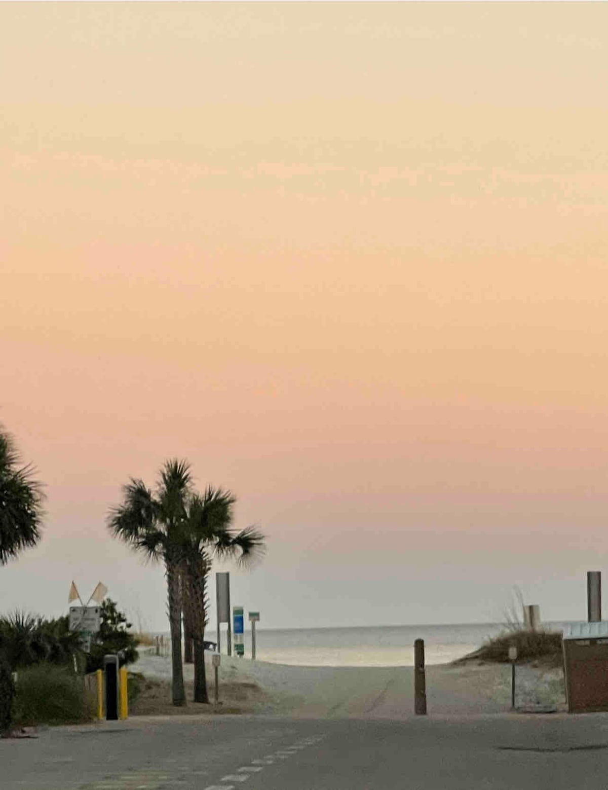 A serene beach pathway is visible, bordered by palm trees and soft vegetation. The horizon displays a gradient of pastel colors, transitioning from soft pink to a calm blue above the gentle waves. Beach access signs can be seen in the distance.