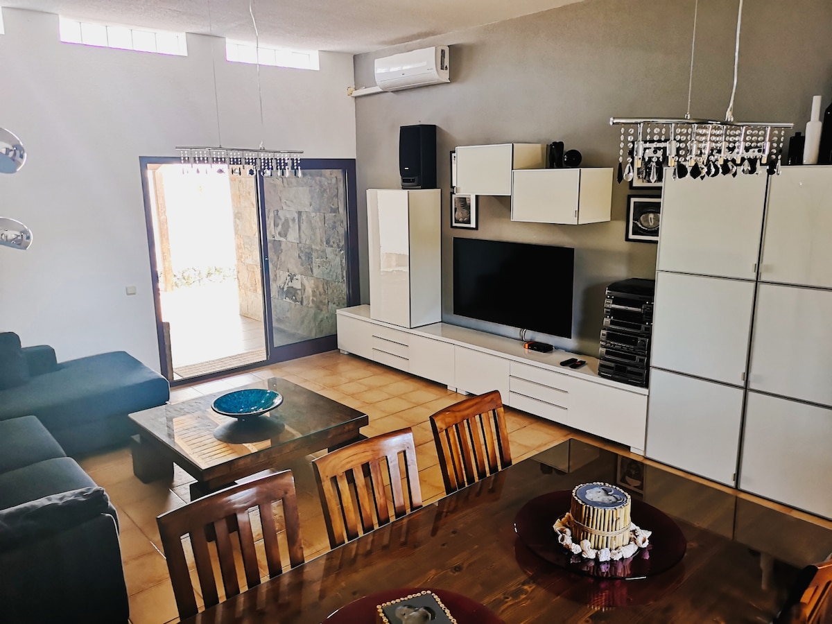 A bright living space features a comfortable sofa and a large dining table set for six. Modern storage units and a flat-screen TV are visible against the neutral walls. An entrance leads to the outside patio, enhancing the connection to the outdoors.