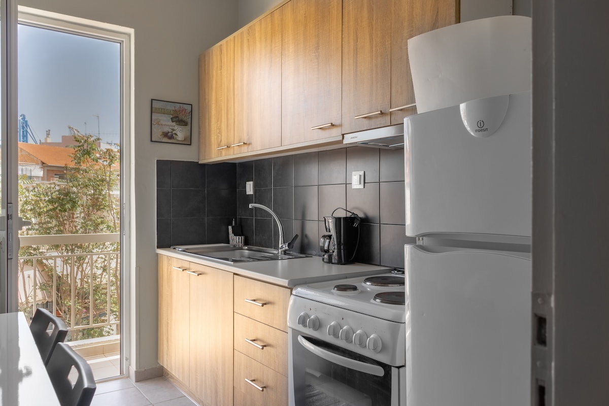 A modern kitchen is shown featuring light wooden cabinetry and gray countertops. Appliances include a white refrigerator, an oven with a stovetop, and a coffee maker. Natural light enters through a large window, illuminating the space. A small dining table with chairs is positioned nearby.
