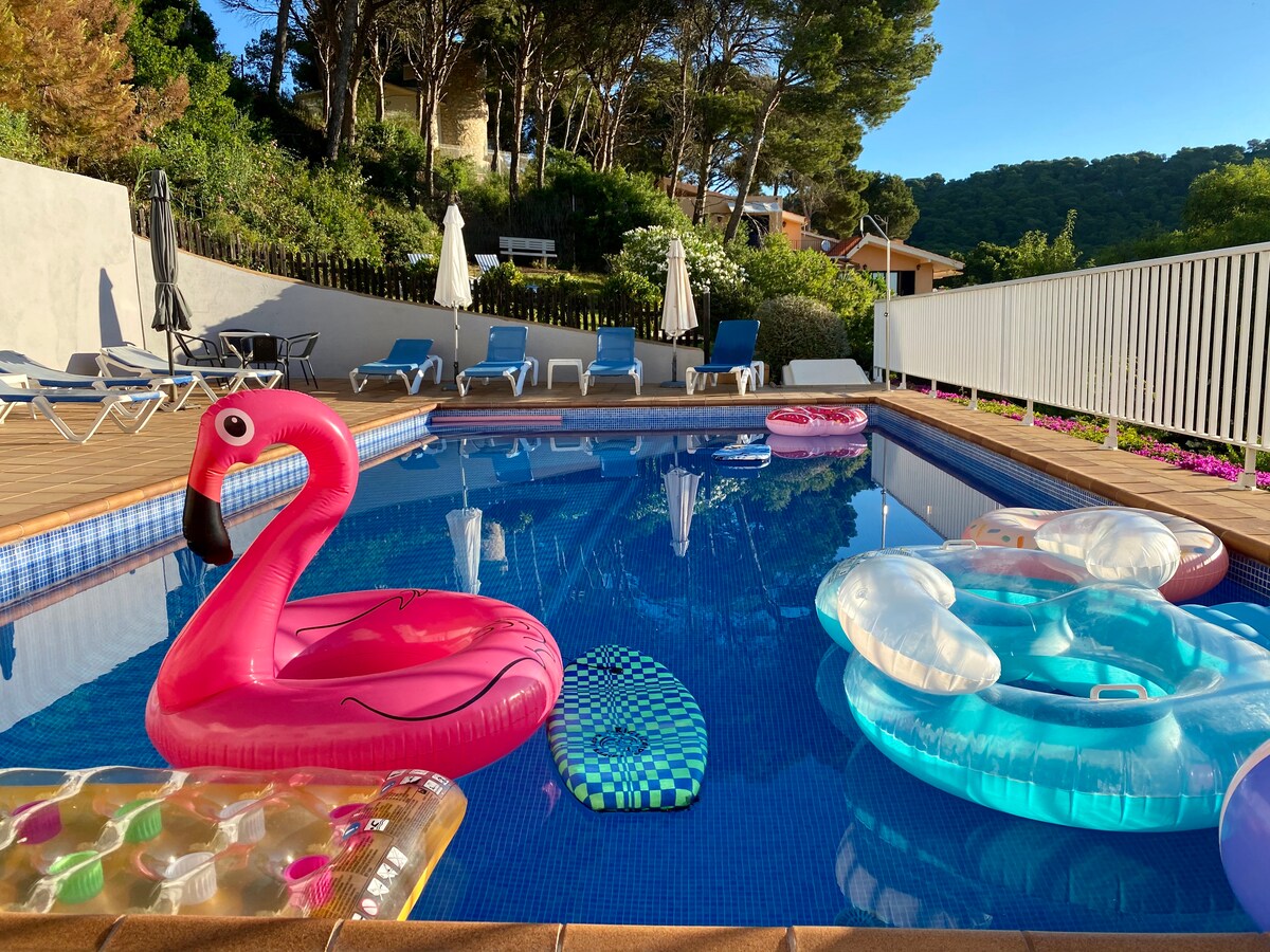 A large swimming pool is surrounded by sun loungers and umbrellas, set against a backdrop of trees. Colorful pool floats in various shapes and sizes float on the water, with a wooden deck providing a space for relaxation.