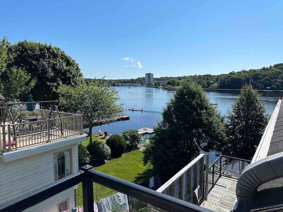A serene view showcases Lake Banook with calm waters reflecting the sky and lush greenery surrounding the area. Kayakers are visible on the water, while nearby lawns are dotted with trees. The balcony railing suggests an elevated perspective of this tranquil setting.