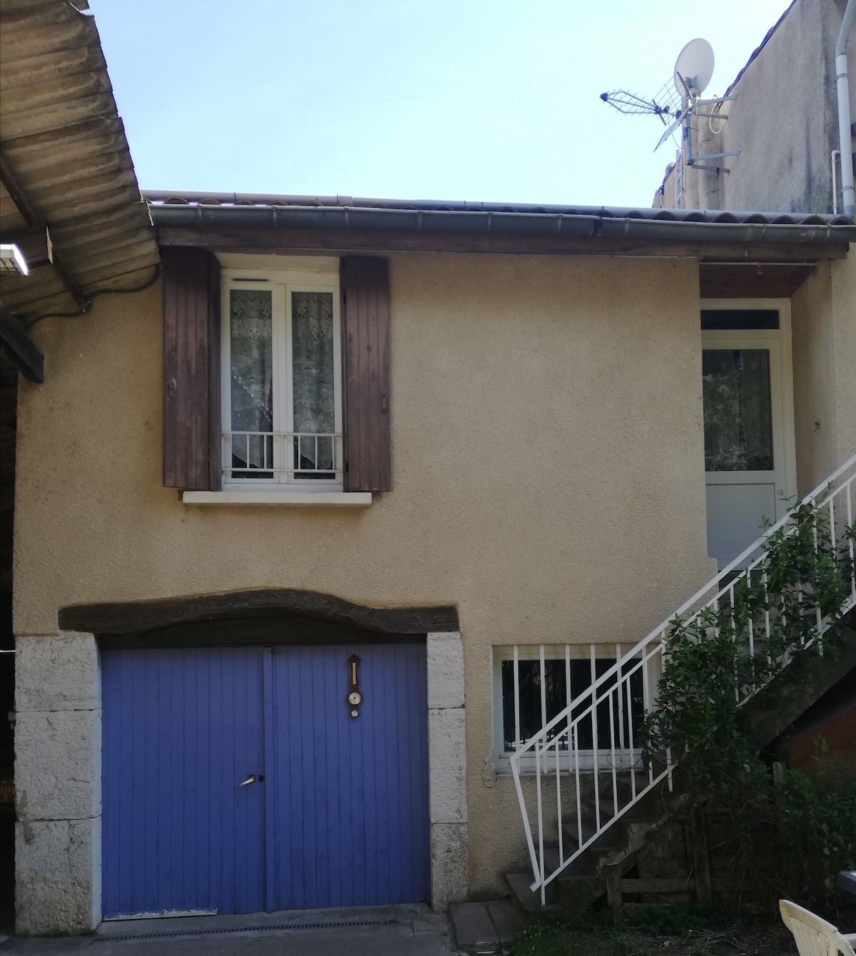 A two-story building is shown with a light brown exterior and decorative wooden shutters in a muted purple hue. A staircase leads to a door on the upper level, while a garage door in a similar shade is located on the lower level. A small tree grows beside the structure.