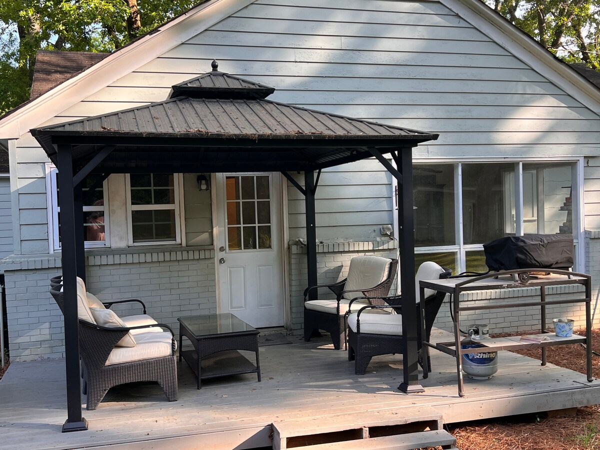 A cozy outdoor space is featured with a gazebo providing shade. Comfortable seating is arranged around a low table, alongside a grill for outdoor cooking. The entrance to the home is visible, with large windows allowing natural light into the interior.