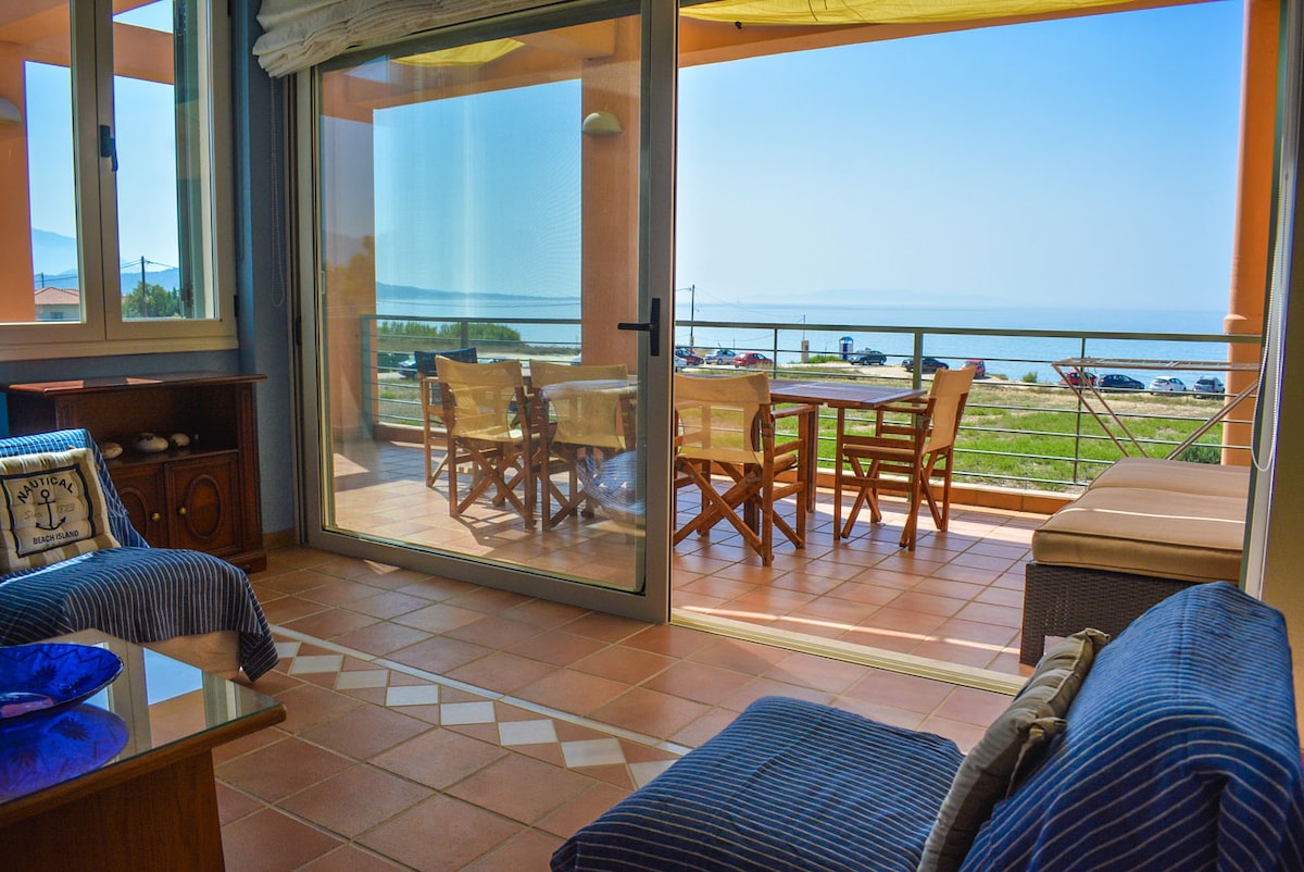 A bright living area features large glass doors opening to a spacious balcony with outdoor dining furniture. The area is illuminated by natural light from the patio, which offers views of the surrounding landscape and sea. Comfortable seating is arranged indoors with decorative accents.