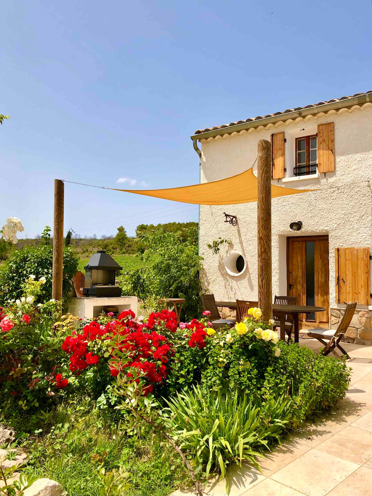 A charming exterior view of the villa is showcased, featuring a patio with a shaded seating area under a sailcloth. Lush gardens filled with vibrant red and yellow flowers surround the space, and a barbecue station adds to the outdoor dining experience.